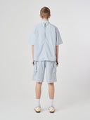 Full-length back view of a male model wearing an Uskees pale blue-grey short-sleeve button front trek shirt paired with matching Uskees technical shorts.