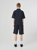 Full-length back view of a male model wearing an Uskees black short-sleeve button front trek shirt paired with matching Uskees technical shorts.