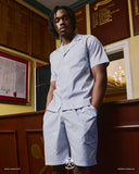 Top-half view of model wearing an Uskees blue, gold and white striped shirt and matching shorts. Hands in deep front pockets.