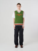 Front shot of male model wearing a green sleeveless sweater with 'USKEES' branding, paired with navy pants.