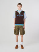 Front shot of male model wearing a brown sleeveless sweater with 'USKEES' branding, light blue shirt, green cargo shorts, and brown shoes on a white background.