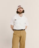 Man wearing an Uskees ecru short sleeve shirt, beige pants, and a cap with 'USK' logo on a beige background.